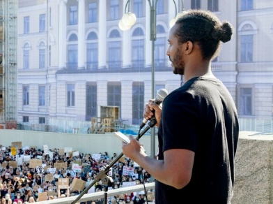 Black lives matter - Liban Sheikh, vas, seisoo ihmisjoukon edessä pitämässä puhetta Senaatintorilla..jpg